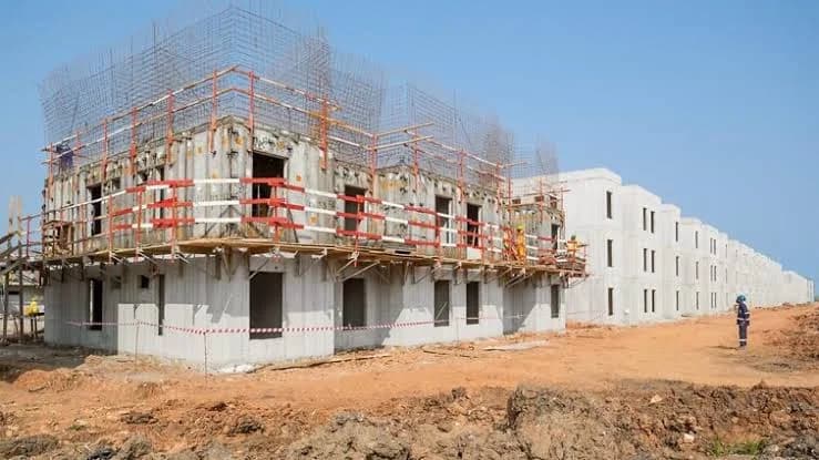 A wide-angle photograph of a large-scale housing construction site featuring multi-story concrete structures in various stages of development. The foreground shows a building with extensive scaffolding and red safety railings, while a long row of completed shells stretches into the distance under a clear blue sky.