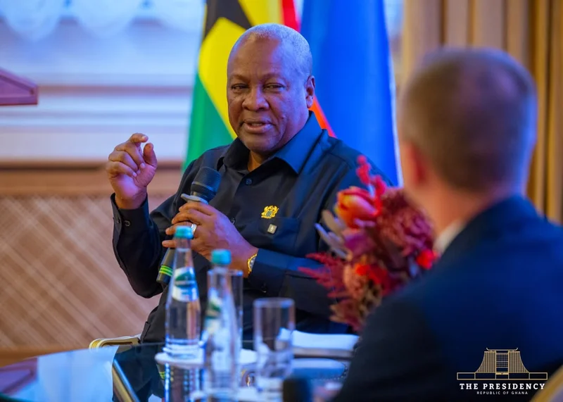 President John Dramani Mahama is pictured speaking into a microphone during an official diplomatic meeting. He is seated at a table with a person in the foreground, with the Ghanaian flag and another international flag visible in the background.
