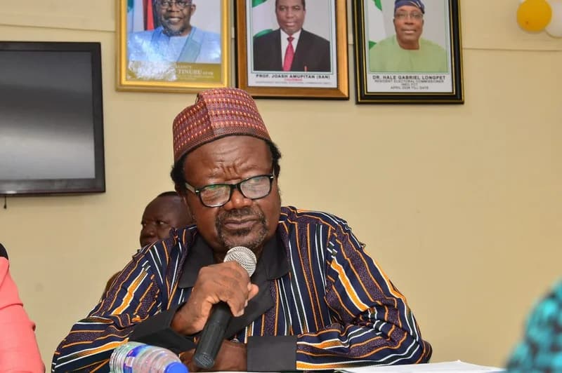 A man wearing a traditional cap and a striped smock is captured speaking into a microphone during an official meeting. Behind him, three framed portraits are displayed on the wall, including Nigerian President Bola Tinubu and officials from the Independent National Electoral Commission (INEC).
