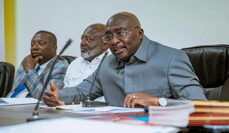 Dr. Mahamudu Bawumia is seen speaking into a microphone during a formal meeting, gesturing with his hand to emphasize a point. He is seated at a conference table with documents and pens, accompanied by two other men in the background.
