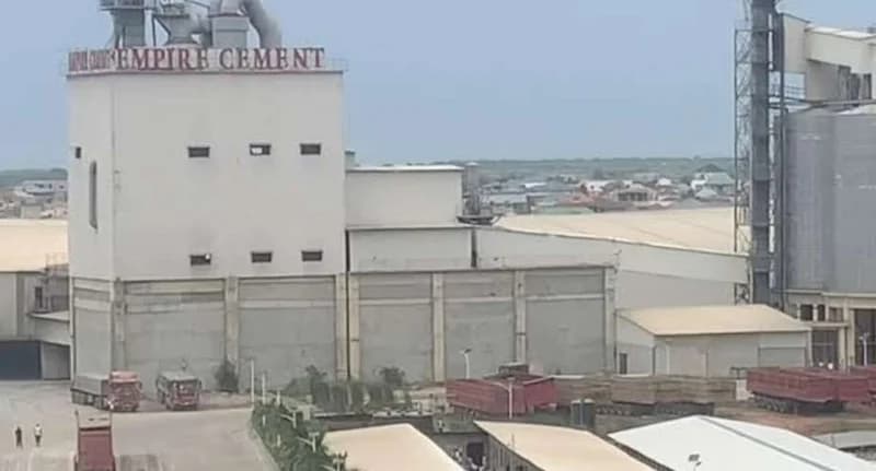 The image shows the industrial facility of Empire Cement, featuring a large white building with the company's name in red text at the top. Several trucks and trailers are parked in the foreground, and large silos and industrial structures are visible under a cloudy sky.