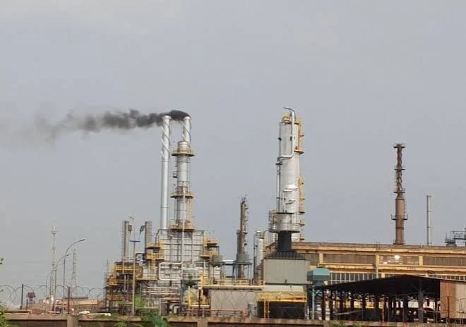 A photograph of the Tema Oil Refinery (TOR) showing industrial towers and processing equipment. Dark smoke is visible billowing from one of the exhaust stacks against a cloudy grey sky.