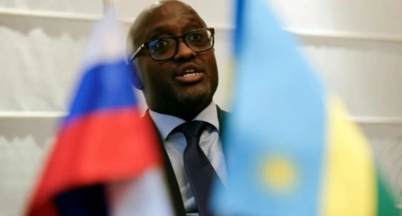 A man wearing glasses and a dark suit with a blue tie is captured mid-speech during an official engagement. He is framed by two blurred flags, identifying as the Russian and Rwandan national flags, indicating a high-level diplomatic setting.