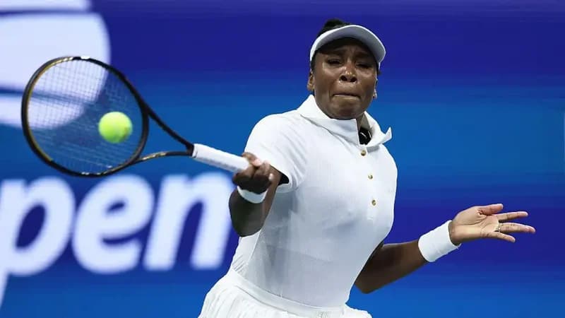 Tennis legend Venus Williams is captured in mid-action during a match at the US Open. She is wearing a white collared shirt, matching visor, and wristbands while swinging her racket at a bright yellow tennis ball against a blue background.