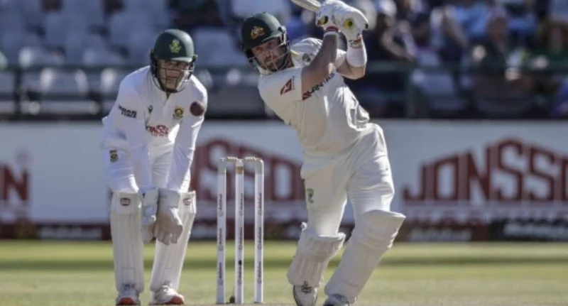 An Australian batsman plays a powerful shot during a Test match against South Africa. A South African wicket-keeper stands behind the stumps watching the play in an outdoor stadium setting.
