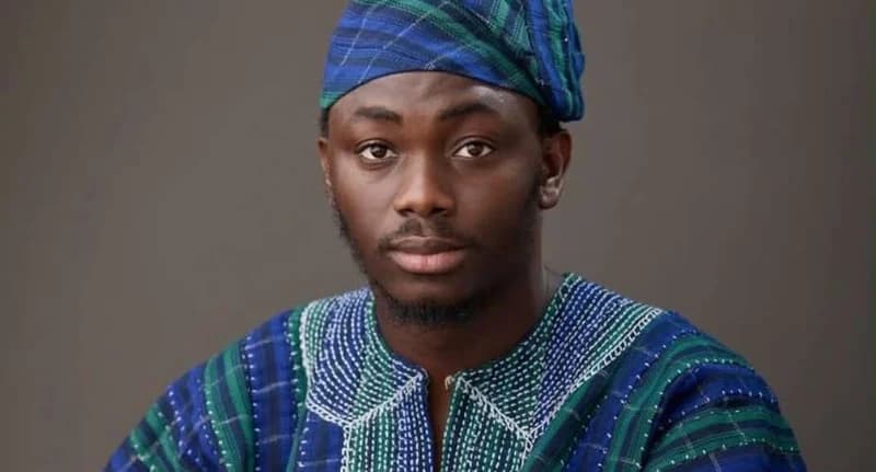A studio portrait of Ewura Adams Karim, a candidate for the NDC Deputy Communications position. He is dressed in a traditional blue and green patterned fugu (smock) with a matching cap, set against a plain grey background.