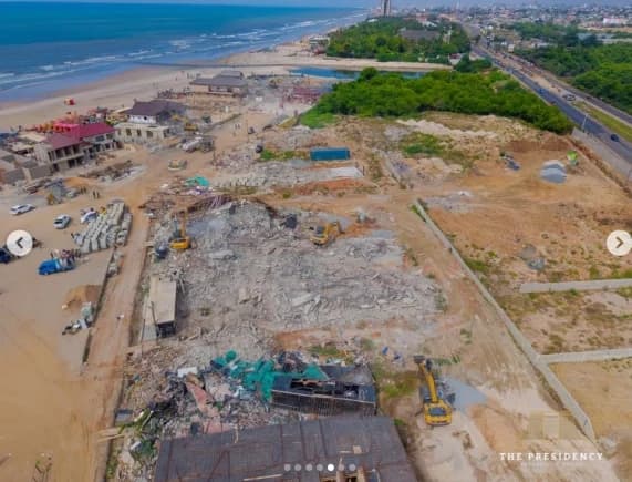 An aerial view shows heavy machinery and excavators amidst the rubble of demolished structures along the coastline at La Pleasure Beach in Ghana. The scene captures the significant scale of the demolition, with debris scattered across the sandy terrain adjacent to the ocean and a nearby highway.