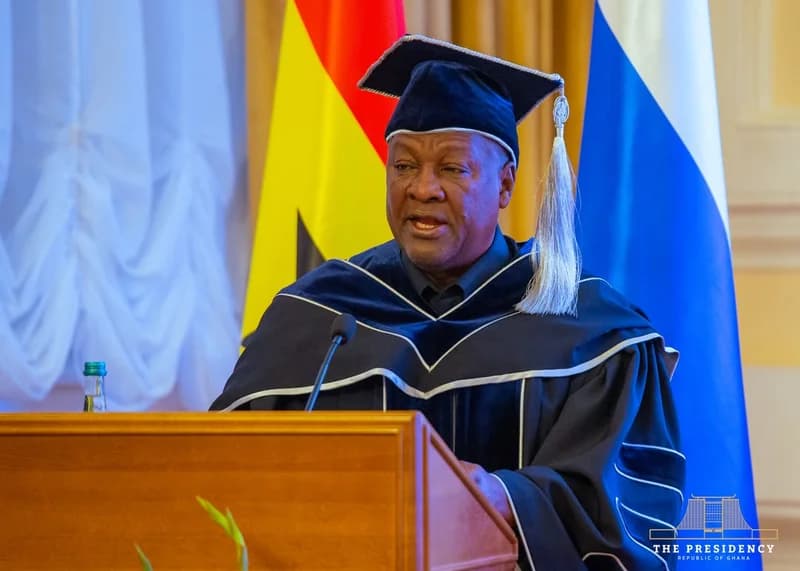 President John Mahama of Ghana is pictured delivering an address from a wooden podium while dressed in formal academic regalia. The scene includes the Ghanaian flag in the background, signifying an official or state-related educational ceremony.