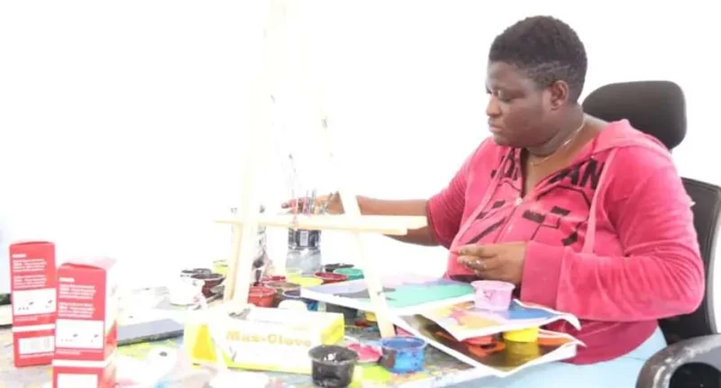 A photograph of a woman, identified as the late policewoman who attempted a paint-a-thon record, sitting at a table filled with art supplies. She is wearing a pink Jordan-branded hoodie and appears focused on her painting, with an easel and numerous paint pots visible in front of her.