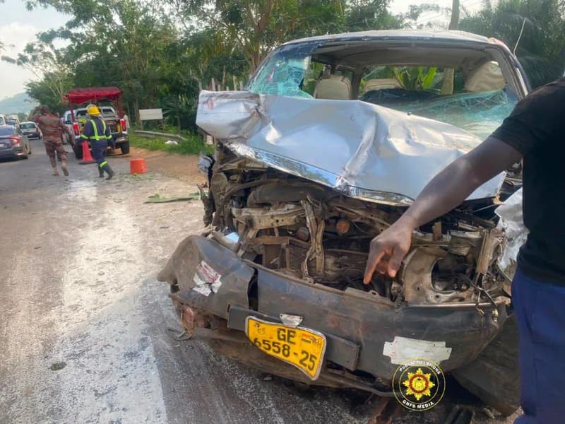 A heavily damaged silver vehicle sits on the side of a road following a collision on the Akuse-Akosombo highway. Personnel from the Ghana National Fire Service are visible in the background attending to the emergency scene.