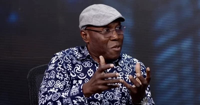 Inusah Fuseini, a prominent Ghanaian politician, is pictured during an interview or presentation, gesturing with his hands while speaking. He is wearing a distinctive blue and white shirt featuring various Adinkra symbols and a grey flat cap.