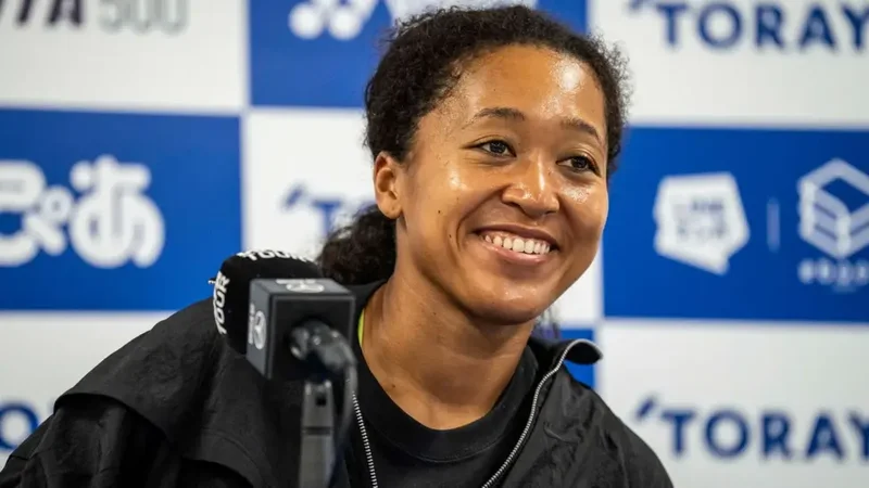 Professional tennis player Naomi Osaka is captured smiling during a press conference. She is seated in front of a blue and white branded backdrop featuring logos for the Toray Pan Pacific Open and the WTA Tour.