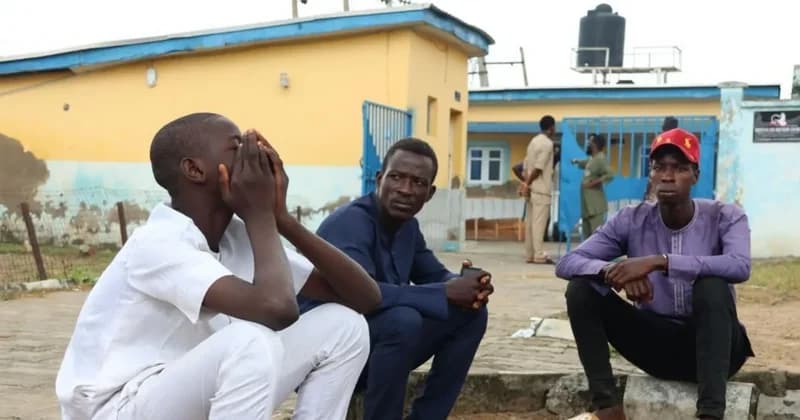 Three young men are pictured sitting outside a yellow building, with one individual holding his head in his hands in a gesture of deep distress. The somber atmosphere and the facility in the background, which features a blue gate and security fencing, suggest a scene related to the Nigerian justice or detention system.