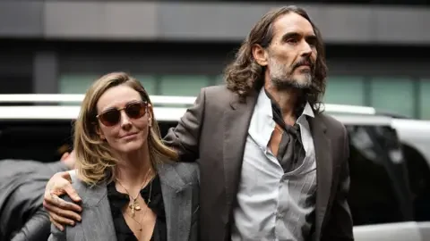 Press Association Russell Brand walking in to court with one arm around his wife's shoulder, she has a grey suit jacket on while he is earing a brown suit jacket and matching cravat