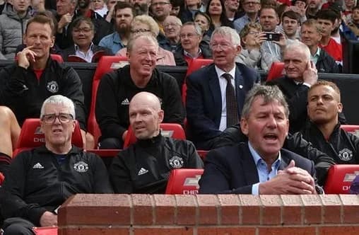 Former Manchester United manager Sir Alex Ferguson and club legend Bryan Robson are seen in the stands alongside coaching staff during a match at Old Trafford. The group appears focused and concerned, reflecting the somber mood often associated with the club's recent performance struggles and structural issues.