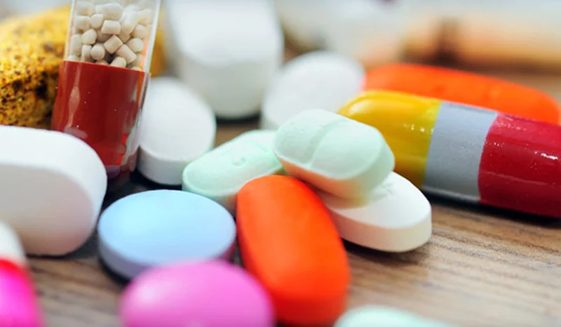 A close-up photograph showcasing a variety of pharmaceutical pills, tablets, and capsules in different colors and shapes. The image features a shallow depth of field, focusing on a collection of medications including an orange oval pill and a green tablet, representing the pharmaceutical industry.