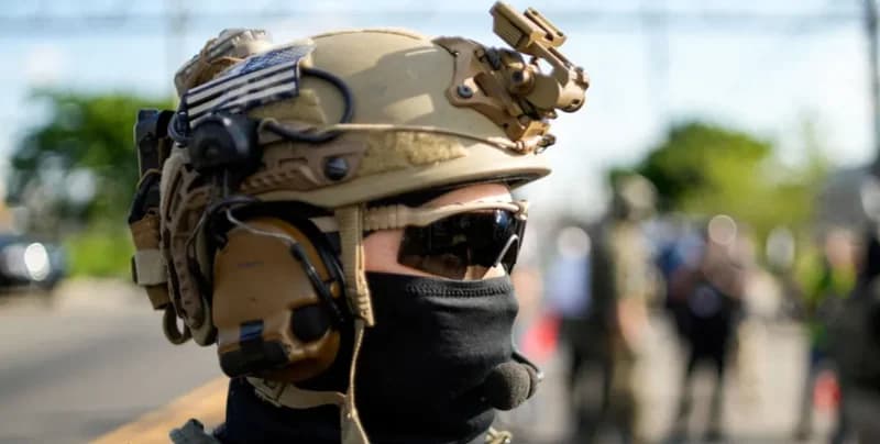 A close-up profile shot of a tactical officer wearing a tan ballistic helmet with a US flag patch, protective sunglasses, and a black face mask. The individual is equipped with professional communication headsets and gear, standing in an outdoor setting with a blurred background.