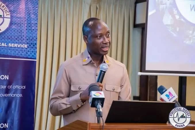 Professor Samuel Kobina Annim, Ghana's Government Statistician, is captured speaking at a podium during a formal event. He holds a microphone and addresses an audience, with multiple media microphones and a Ghana Statistical Service banner visible in the background.