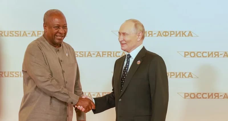 President John Dramani Mahama of Ghana shares a handshake with Russian President Vladimir Putin during a formal diplomatic encounter. The leaders are positioned in front of a backdrop branded with "Russia-Africa" in English and Cyrillic, reflecting talks on international cooperation and trade.