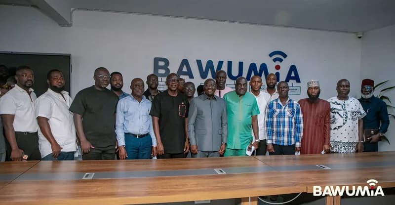 Dr. Mahamudu Bawumia is pictured standing in the center of a group of men in a formal meeting room setting. They are gathered behind a large wooden conference table in front of a wall prominently displaying the 'BAWUMiA' campaign branding.