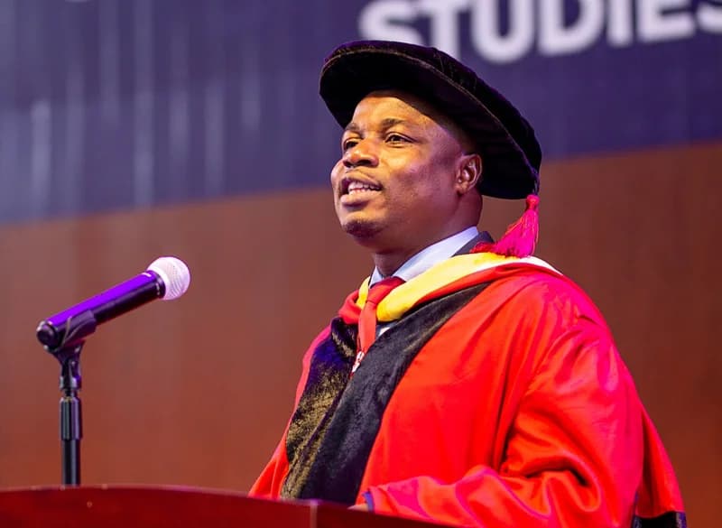 Professor Abednego Feehi Okoe Amartey, Vice-Chancellor of the University of Professional Studies, Accra (UPSA), delivers a speech at a graduation ceremony. He is seen wearing traditional red and black academic regalia while standing behind a podium with a microphone.