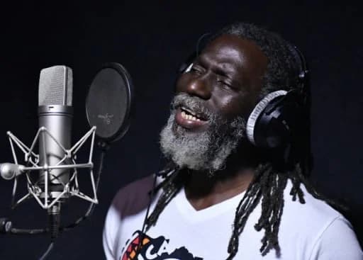 Ivorian reggae star Tiken Jah Fakoly is captured singing into a studio microphone with intense focus. He is wearing professional over-ear headphones and a white t-shirt featuring a map of Africa, set against a dark, minimalist studio background.