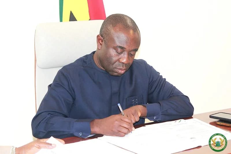 A photograph showing Ghanaian Minister for Employment and Labour Relations, Ignatius Baffour-Awuah, signing a document at a desk. Behind him is a portion of the Ghanaian flag, and the official seal of the ministry is visible in the bottom right corner.