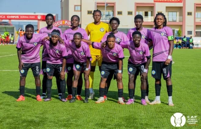 A team photograph of Ampem Darkoa Ladies FC players posing on a football field in their pink and black match kits. The eleven players are arranged in two rows, with the goalkeeper in yellow at the center, set against a backdrop of stadium facilities and the CAF Women's Champions League logo.