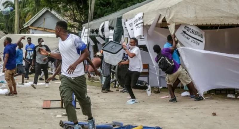 A chaotic scene at a Tanzanian polling station showing people running and disrupting the voting process. Signs indicating a 'POLLING STATION' are visible on white tents as individuals appear to be carrying away ballot boxes amidst the confusion.