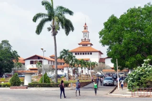 A view of the iconic Balme Library clock tower at the University of Ghana, Legon. Students are seen walking across the campus foreground with lush greenery and palm trees surrounding the white buildings with orange-tiled roofs.