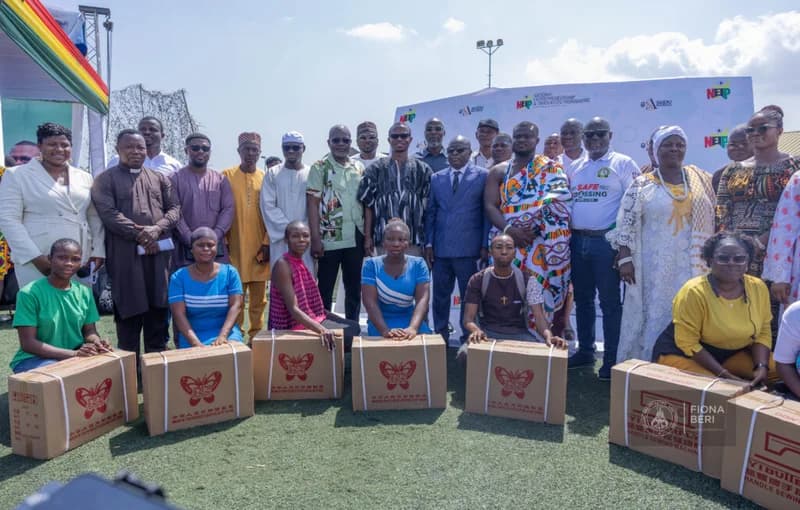 A large group of Ghanaian officials, traditional leaders, and beneficiaries gather for an official distribution ceremony outdoors. In the foreground, several individuals pose with boxed sewing machines featuring the Butterfly brand logo, while a large backdrop behind them displays the National Entrepreneurship & Innovation Programme (NEIP) branding.