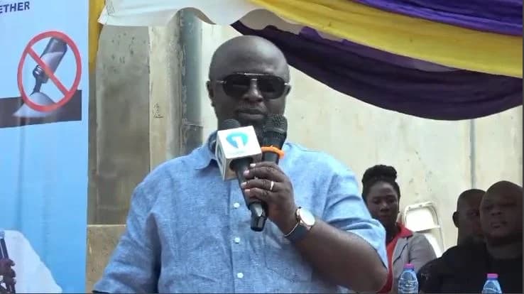 Sam Pyne, the Mayor of Kumasi, is captured speaking into two microphones during a public gathering. He is wearing a light blue button-down shirt and dark sunglasses, standing under a yellow and purple decorative canopy with a banner partially visible to his left.