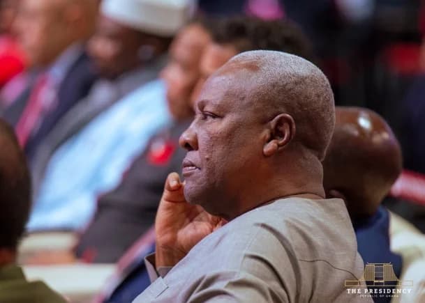 President John Dramani Mahama is captured in a profile view while attending a formal event, appearing to listen attentively to proceedings. He is seated among other dignitaries in a brightly lit conference or summit setting, with an official watermark in the bottom corner.