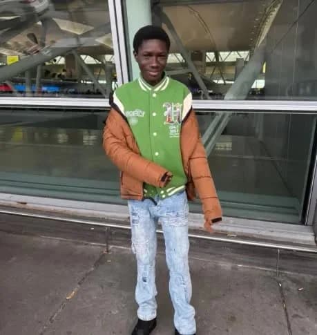 A photograph shows a young teenage boy of Ghanaian descent posing in front of a modern glass-walled building. He is wearing a green and white varsity jacket, a brown puffer jacket slung over his arms, and light-colored ripped jeans.
