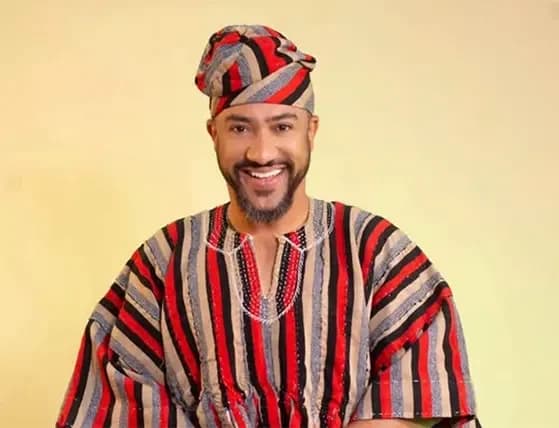 A studio portrait of Ghanaian actor Majid Michel smiling warmly. He is wearing a traditional Northern Ghanaian smock (fugu) with vertical red, black, and beige stripes, paired with a matching traditional cap.