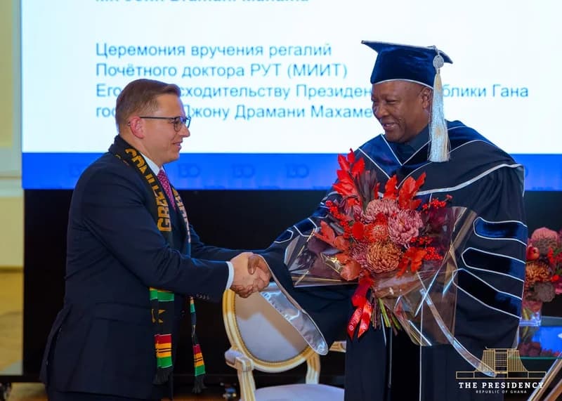 President John Dramani Mahama of Ghana is shown shaking hands with a Russian official during a ceremony where he was awarded an honorary doctorate. Mahama is dressed in blue academic regalia and holding a large bouquet of red and orange flowers, standing in front of a screen displaying Russian text about the event.