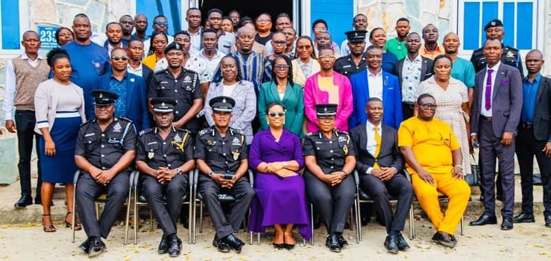 A group photograph featuring high-ranking officers of the Ghana Police Service and civilian officials gathered for a formal engagement. The participants are arranged in multiple rows, with seated senior officers in black uniforms in the front and various personnel standing behind against a light-colored building.