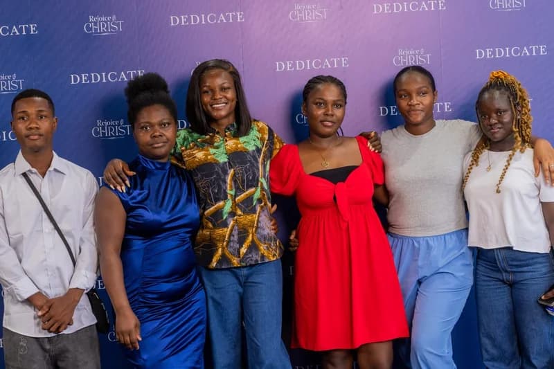 A group of six young people, including five women and one man, are posing together in front of a blue promotional backdrop. The individuals are dressed in various casual and semi-formal outfits, smiling for the camera at what appears to be a film event in Accra.