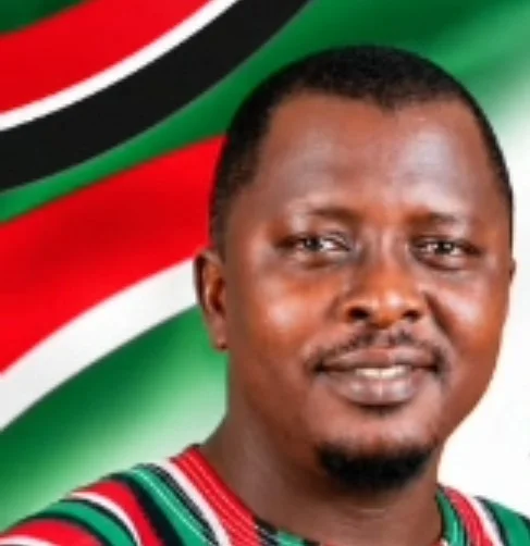 A portrait of a man, identified as the Assin Central NDC Chairman, smiling against a background featuring the colors of the National Democratic Congress. He is wearing a smock with a pattern consisting of red, green, black, and white stripes, which are the official colors of the NDC party in Ghana.