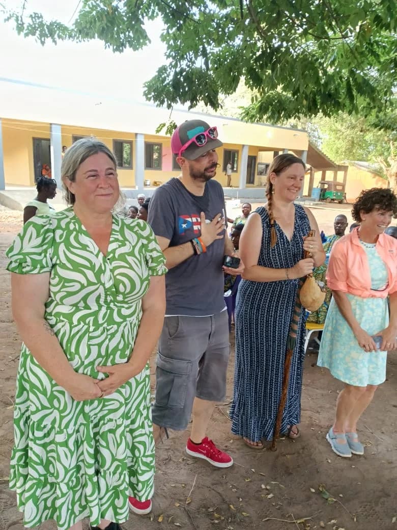 A group of four visitors stands in an outdoor courtyard in front of a yellow building, likely a school or community center. One woman is holding a traditional staff topped with a gourd, suggesting an engagement with local cultural practices or a storytelling ceremony as referenced in the article context.