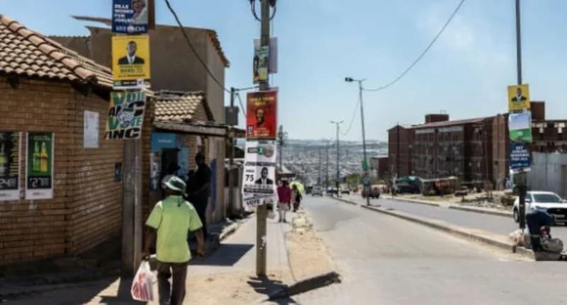 A street scene in a South African urban area or township showing utility poles covered in various political campaign posters. A man in a lime green shirt walks past brick buildings while several other pedestrians and vehicles are visible in the background under a bright sky.