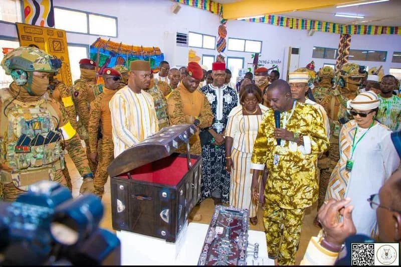 The image shows Burkinabe President Captain Ibrahim Traoré and other dignitaries observing a display at an exhibition. A man in a gold-patterned traditional outfit is speaking into a microphone while showcasing an open wooden chest containing artisanal items, while armed security personnel stand guard.