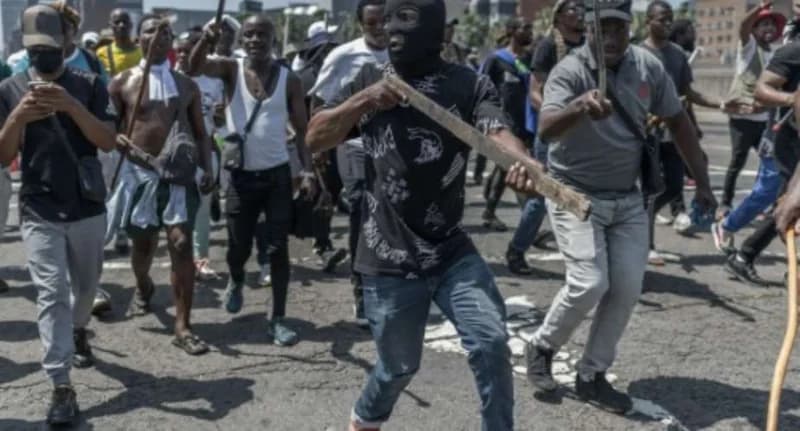 A group of men are seen marching through a street during a protest, with some individuals brandishing wooden sticks. A man in the center foreground wears a black balaclava, emphasizing the tense and potentially confrontational atmosphere of the demonstration in an urban setting.