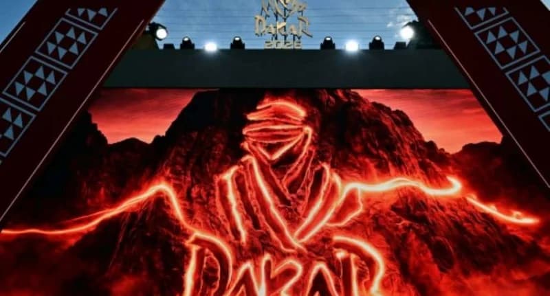 A large digital display features the glowing red logo of the Dakar Rally, showing the iconic Tuareg head against a mountainous landscape. The stage is framed by decorative pillars and illuminated by spotlights, used for an official event announcement.