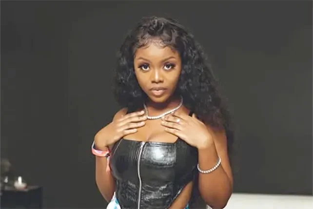 A studio portrait of Ghanaian singer Gyakie looking directly at the camera. She is wearing a black zippered top, silver-toned jewelry including a necklace and bracelet, and her hair is styled in long waves.