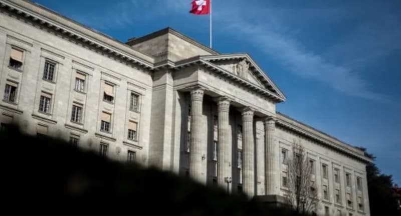 A photograph of the exterior of the Federal Supreme Court of Switzerland, featuring classical architecture with large stone columns. The Swiss national flag flies prominently at the top of the building against a clear blue sky.