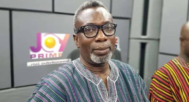 A portrait of veteran Ghanaian highlife musician K.K. Kabobo posing in a media studio. He is wearing black-rimmed glasses and a traditional striped smock (fugu) with the Joy Prime television logo visible in the background.