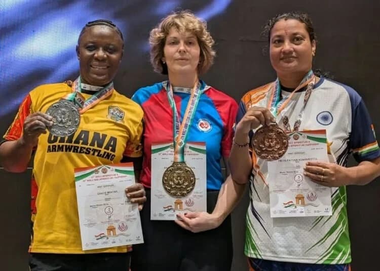 Ghanaian armwrestler Grace Minta stands on the podium alongside two other competitors at the World Armwrestling Championship. She is visible on the left wearing a yellow jersey with 'GHANA ARMWRESTLING' printed on it, holding a silver medal and an official achievement certificate.