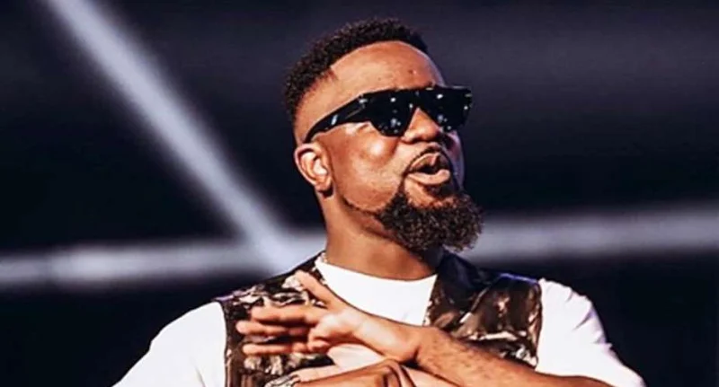 A close-up photograph of Ghanaian rapper Sarkodie during a performance or public appearance. He is seen wearing dark sunglasses, a white t-shirt, and a patterned vest while gesturing with his hands.