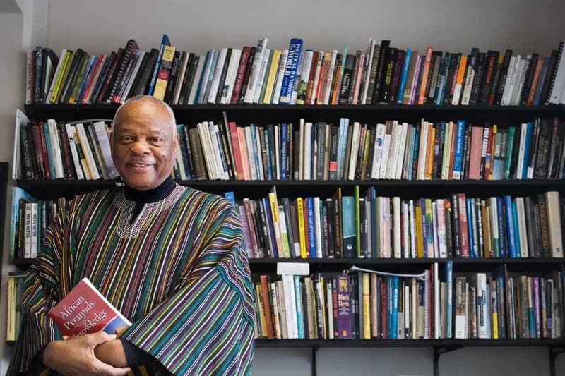A portrait of Professor Molefi Kete Asante smiling while holding his book, 'African Pyramids of Knowledge.' He is standing in front of large bookshelves filled with academic texts and wearing a traditional striped African garment.